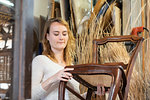 Young female basket maker examining woven seat on dining chair in workshop