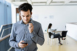 Businessman using smartphone in office corridor, colleague reading in background