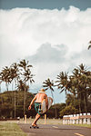 Mid adult male skateboarder carrying surfboard, skateboarding on rural road, rear view, Haiku, Hawaii, USA