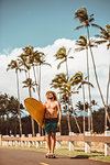 Mid adult male skateboarder wearing straw hat, standing on skateboard carrying surfboard on rural road, portrait, Haiku, Hawaii, USA