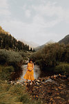 Pregnant woman in orange maxi dress crossing stepping stones in rural river, Mineral King, California, USA