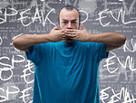 Conceptual front view portrait of a man covering his mouth with his hands while looking at camera against gray background with speak no evil message