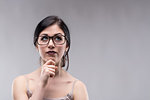 Attractive woman wearing glasses deep in thought looking up with a serious pensive expression and her hand to her chin over a grey background with copy space