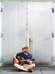 Photo of a young adult male smoking a cigar and playing an acoustic guitar on the street.