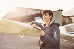 Smiling young woman with camera, she is using a smart phone and leaning on a propeller airplane wing, travel and tourism concept