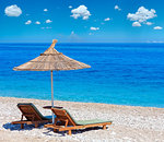 Summer morning pebbly beach with sunbeds and straw sunshade (Albania). Deep blue sky with same cumulus clouds. Two shots stitch high resolution image.