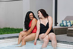 Two mid adult women sitting on outdoor swimming poolside chatting and laughing together, Cape Town, South Africa