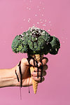 Studio shot of man's hand holding ice cream cone with broccoli on top, dripping chocolate sauce and sprinkles, against pink background