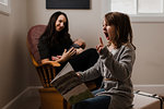 Girl with book pulling faces, while mother cradles baby brother in living room armchair