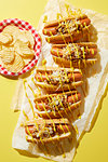 Row of coney hotdogs with drizzled mustard and plate of potato chips on yellow background, overhead view