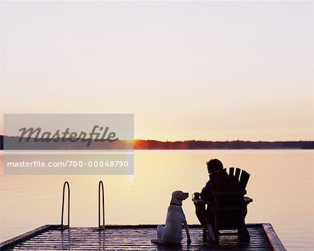 Rückansicht des Mannes im Adirondack Stuhl am Dock mit Hund bei Sonnenuntergang Bala, Ontario, Kanada