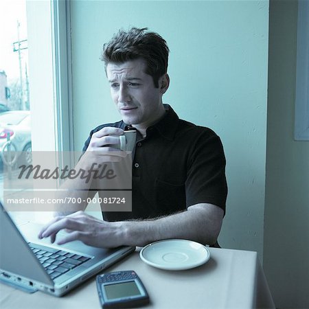 Man Sitting at Table by Window in Cafe, Using Computer, Holding Cup