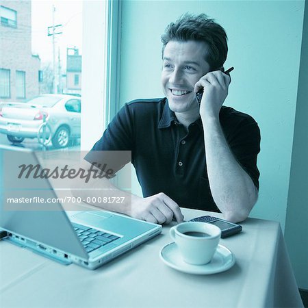 Man Using Cell Phone in Cafe with Laptop Computer on Table