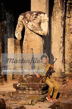 Man Sitting by Statue, Holding Gun at Preah Khan Temple Siem Reap, Cambodia