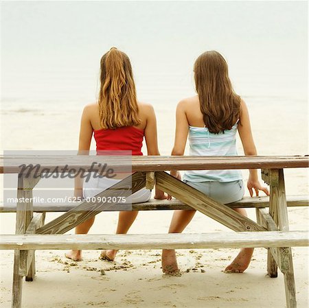 Rückansicht der Mädchen und ein junges Mädchen sitzt auf einem Picknick-Tisch am Strand