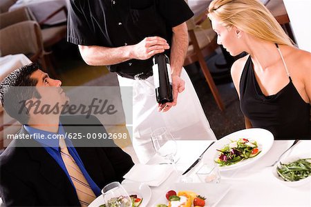 Mid adult man and a young woman sitting at the table with a waiter showing them a wine bottle
