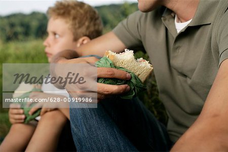 Father and son sitting on meadow while they eat sandwiches