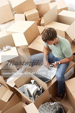 Man Sitting amongst Moving Boxes with Laptop Computer and Cellular Phone