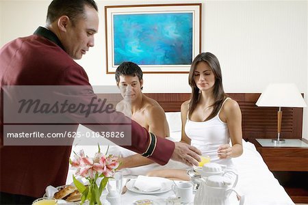 Side profile of a waiter serving a young woman and a mid adult man breakfast in bed