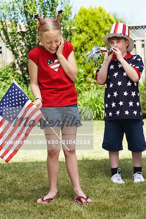 Girl Holding American Flag with Boy Wearing Stars and Stripes Top and Hat, Blowing Noisemaker Horn
