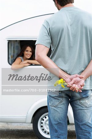 Woman Looking out Trailer Window at Man Holding Flowers Behind Back