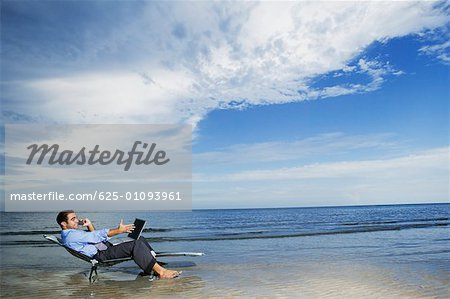 Side profile of a young man sitting on the beach with a laptop