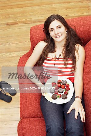 High angle view of a young woman lying on a couch and holding a plate of strawberries with chocolates