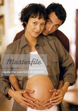 Man touching pregnant woman's stomach from behind, looking at camera, portrait