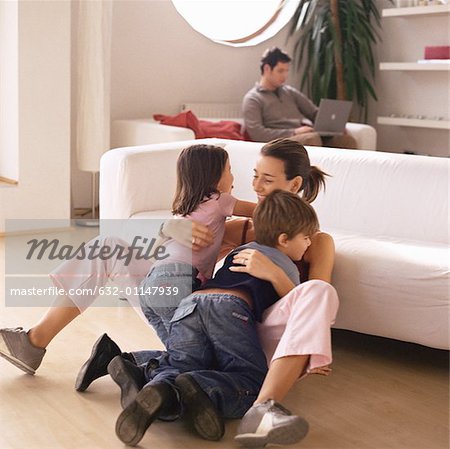 Woman being hugged by two children, man working on laptop in background