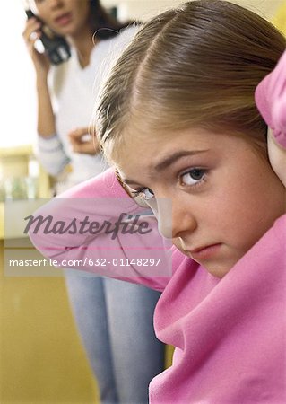 Petite fille avec les mains sur les oreilles, regardant la caméra, close-up, femme debout parlait au téléphone en arrière-plan