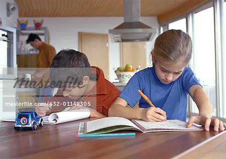 Son and daughter writing at table, father in background cooking, blurred.