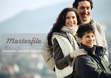Adult man and woman with boy, woman's hand on boy's shoulder, waist up, mountain landscape blurred in background