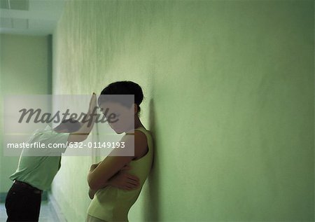 Frau mit dem Rücken gegen die Wand, Arme gefaltet, Mann im Hintergrund deprimiert an die Wand gelehnt