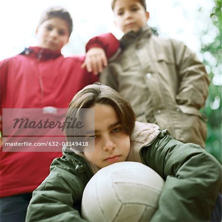 Three boys outside looking at camera, one holding ball, low angle view