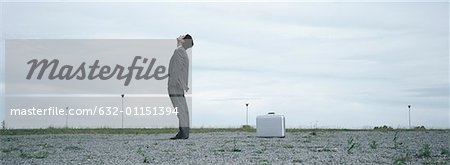 Man standing in abandoned lot looking up at sky, metallic briefcase on ground behind him