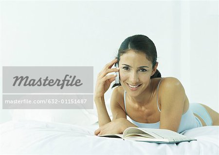 Young woman lying on stomach in underclothes holding cell phone to ear, smiling at camera