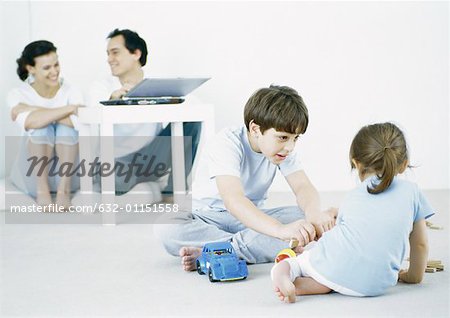 Garçon et fille jouant sur le plancher, parents en arrière-plan à l'aide d'ordinateur portable