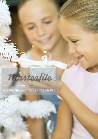 Girl holding present next to christmas tree, sister watching