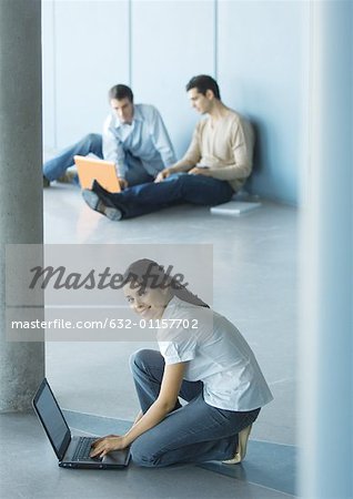 Young woman crouching sitting on floor, using laptop, men sitting in background