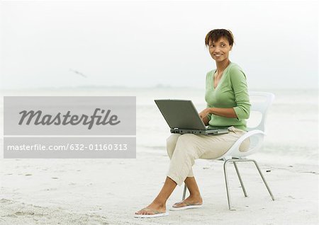 Woman on beach, sitting in chair using laptop