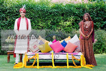 Portrait of a newlywed couple in traditional wedding dress standing near a bed in a lawn