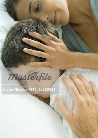 Child lying in bed, parent's hands on head and shoulder