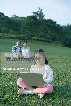 Girl sitting in grass using laptop, parents sitting in background