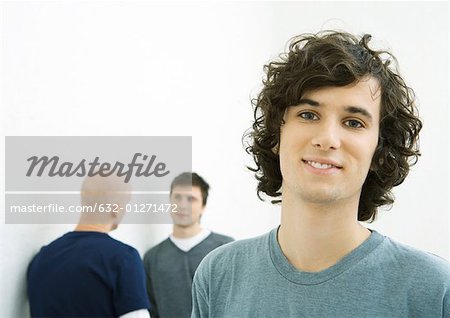 Jeune homme souriant à la caméra, deux jeunes hommes parlant en arrière-plan