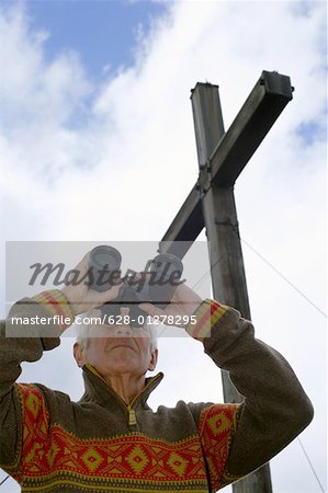 Senior adult man standing under a wooden cross and is looking through binoculars, low angle view