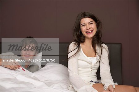 Young brunette woman is sitting at the edge of a bed while her boyfriend is looking at a pregnancy test