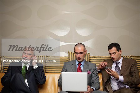 Drei Geschäftsmänner auf einem Sofa mit verschiedenen Kommunikationsgeräten