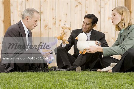 Two businessmen and a businesswoman playing with toys sitting on a lawn