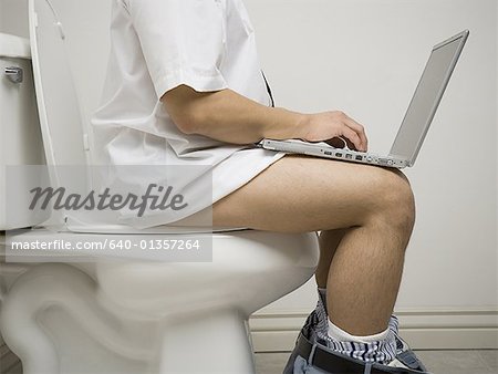 Milieu vue en coupe d'un jeune homme assis sur le siège de toilette à l'aide d'un ordinateur portable