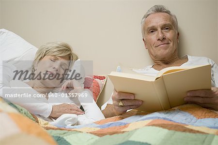 Portrait d'un homme senior allongée sur un lit, tenant un livre pendant qu'une femme senior dort